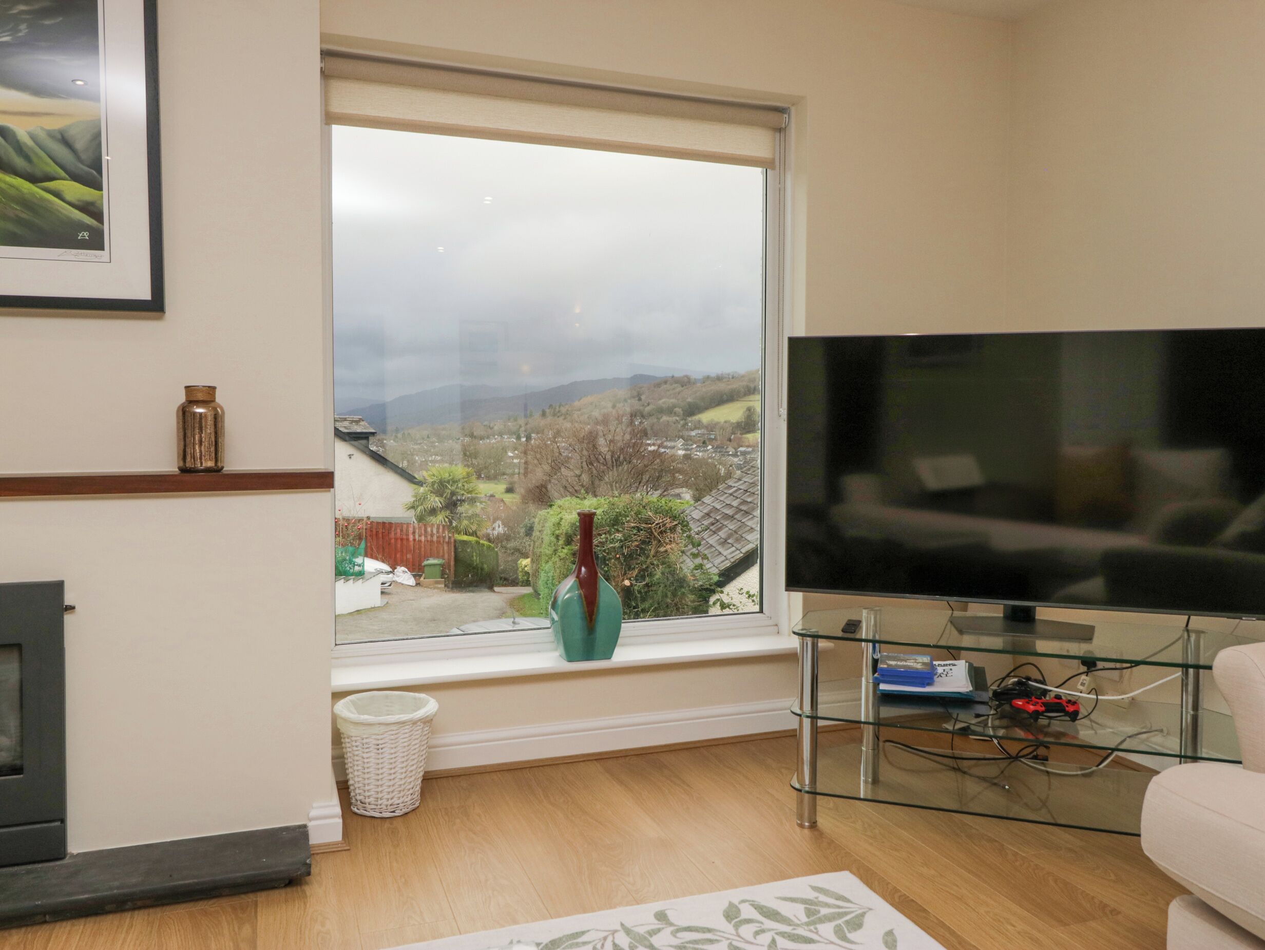 Ein Wohnzimmer mit Fernseher, Kamin, Fenster mit Aussicht und einem Glastisch.