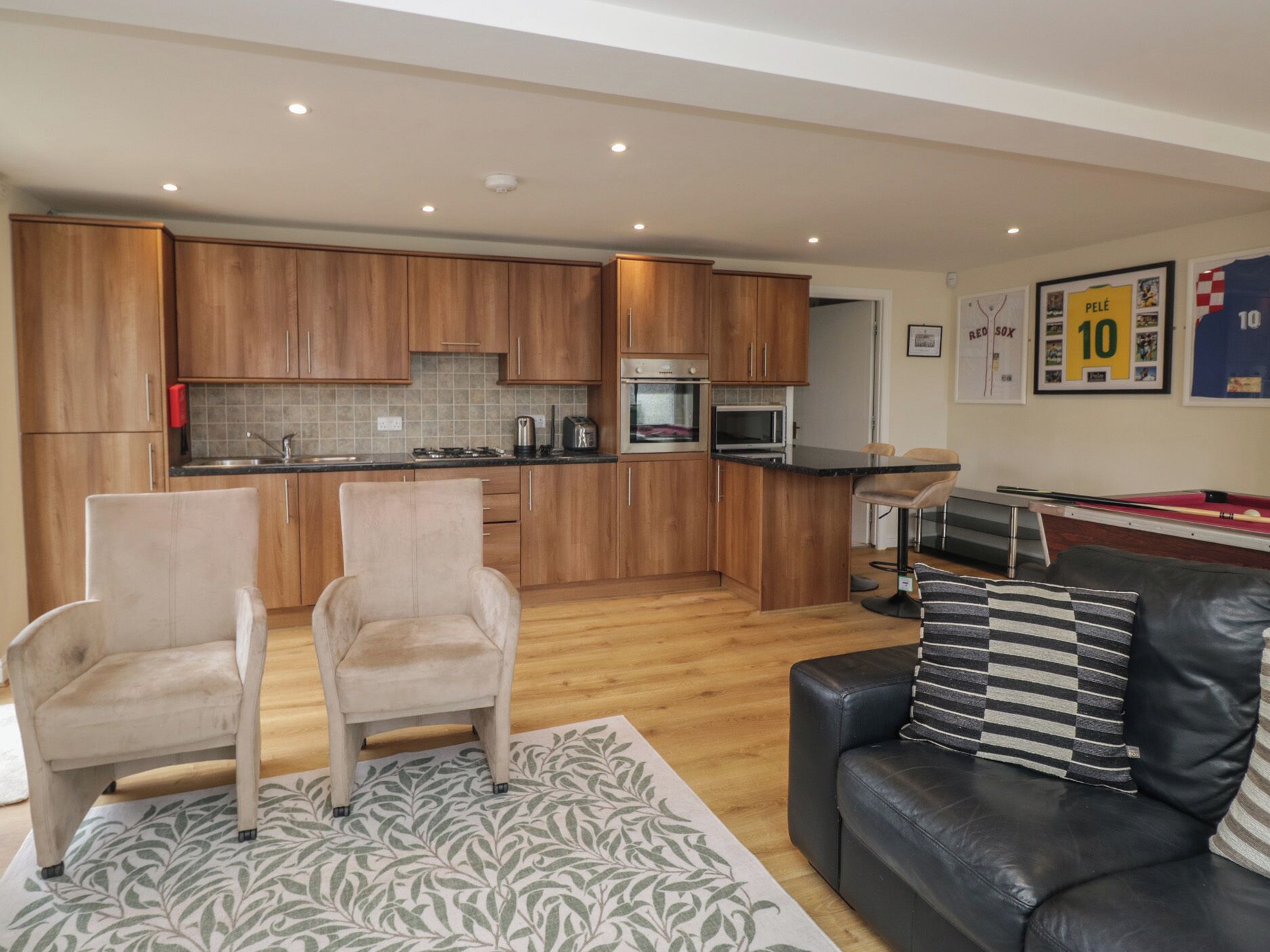 A modern kitchen with wooden cabinets, a dining area with a black table, and a living room with a leather sofa and a patterned rug.