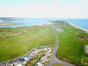 An aerial view of a coastal area with a road leading to a cluster of houses, a large open field, and a beach. - Bayview Cottage (Bournemouth)