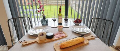 A dining table set with plates, a bottle of wine, a bowl of cherries, a cheese board, and a loaf of bread.