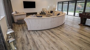 A modern living room with a large sectional sofa, a flat-screen TV mounted on the wall, and a large window offering a view of the outdoors. - 85A Braybrooke Road (Kettering)