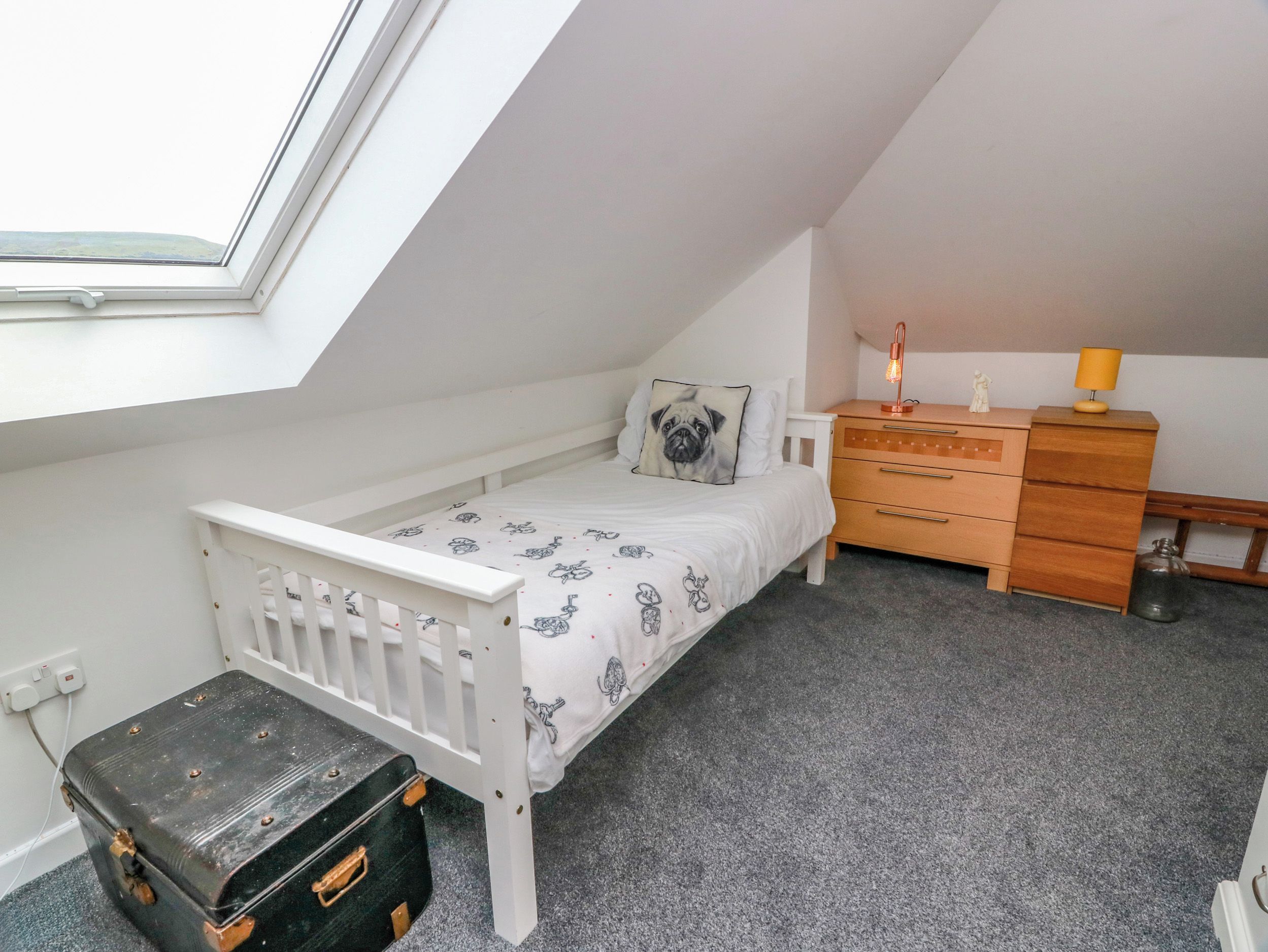 A small, single-bed room with a sloped ceiling, a bedside table with a lamp, a dresser, and a suitcase.