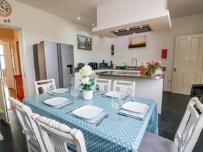 A dining area with a table set for four, a kitchen in the background, and a clock on the wall.