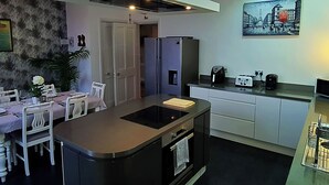 A modern kitchen with a central island, built-in oven, and a refrigerator. There is a dining area with a table and chairs, and a painting on the wall. - Ailsa View (Girvan)