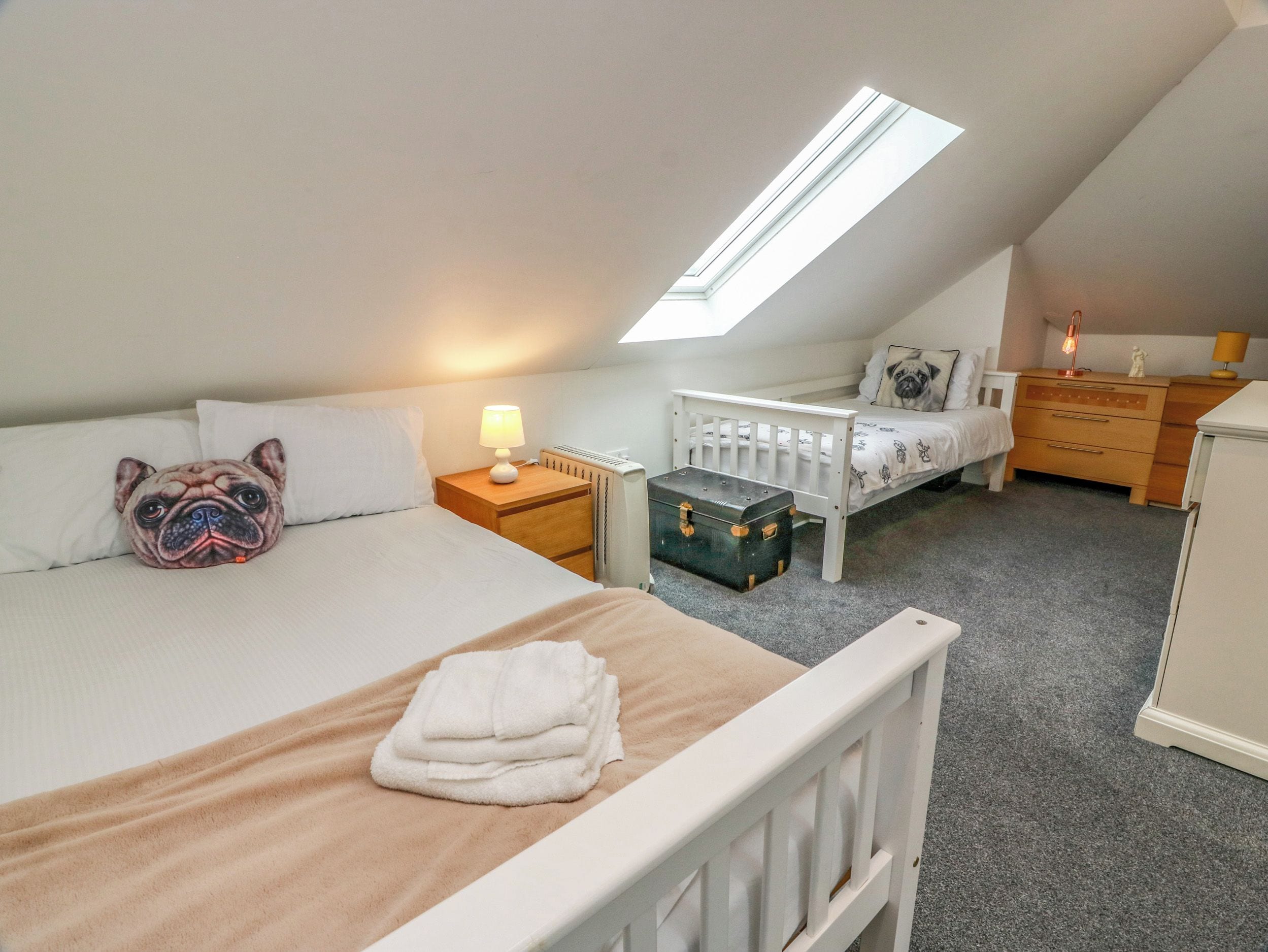 Une chambre avec un plafond en pente, un lit avec un oreiller, une table de chevet avec une lampe, une petite commode, un radiateur et une valise.