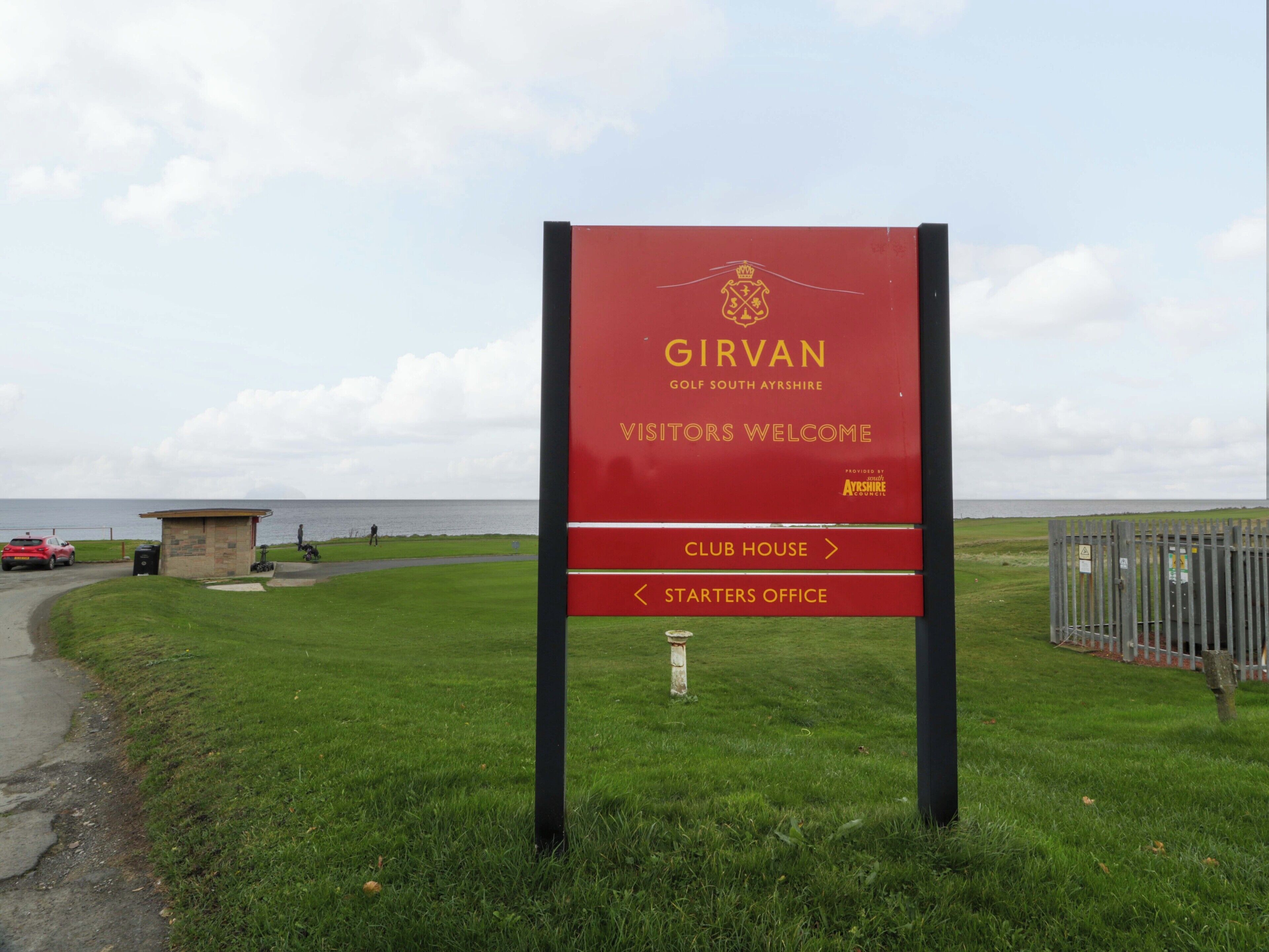 Señal roja con letras doradas para Girvan Golf South Ayrshire, dando la bienvenida a visitantes e indicando el camino a la casa club y la oficina de los iniciadores.