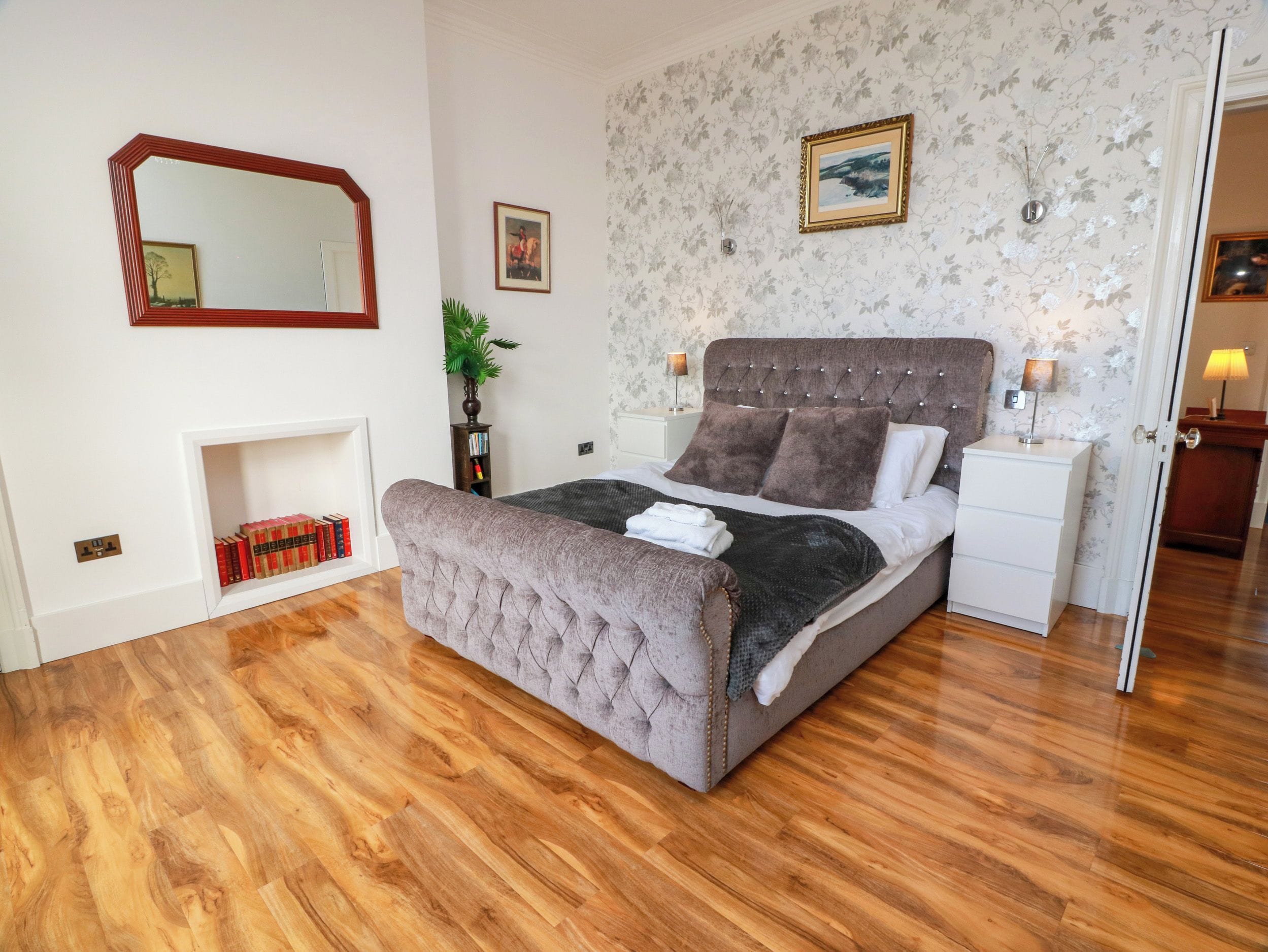 Une chambre à coucher avec un grand lit, une table de chevet, une commode, un miroir, une cheminée et un mur recouvert d’un papier peint à motifs floraux.