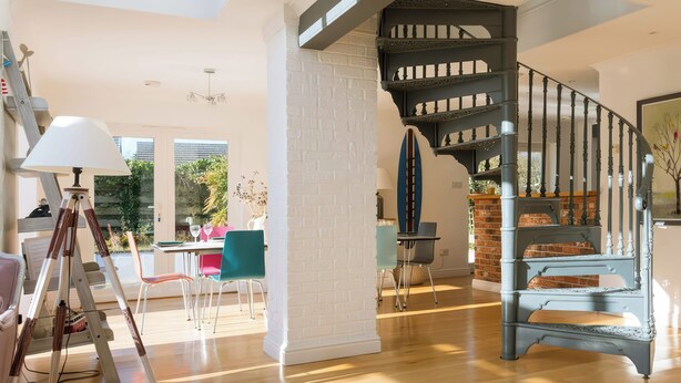 Ein geräumiger Wohnbereich mit Holzboden, einem Esstisch mit Stühlen, einer Dreibeinlampe und einer Treppe mit dekorativer Geländer.