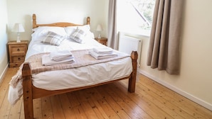 A neatly made bed with white linens and plaid pillows, a wooden headboard, a nightstand with a lamp, and a window with curtains. - Bryn Uchaf (Tyn-y-Gongl)
