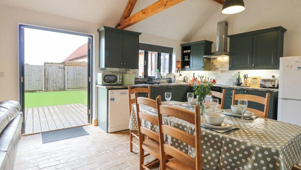 A kitchen with dark cabinets, a microwave, and a refrigerator. There is a dining table with chairs and a vase of flowers. - The Old Dairy (Dereham)