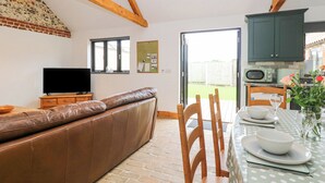 A cozy living room with a leather sofa, a dining table set for two, wooden chairs, a television, and a kitchen area with cabinets and a microwave. - The Old Dairy (Dereham)