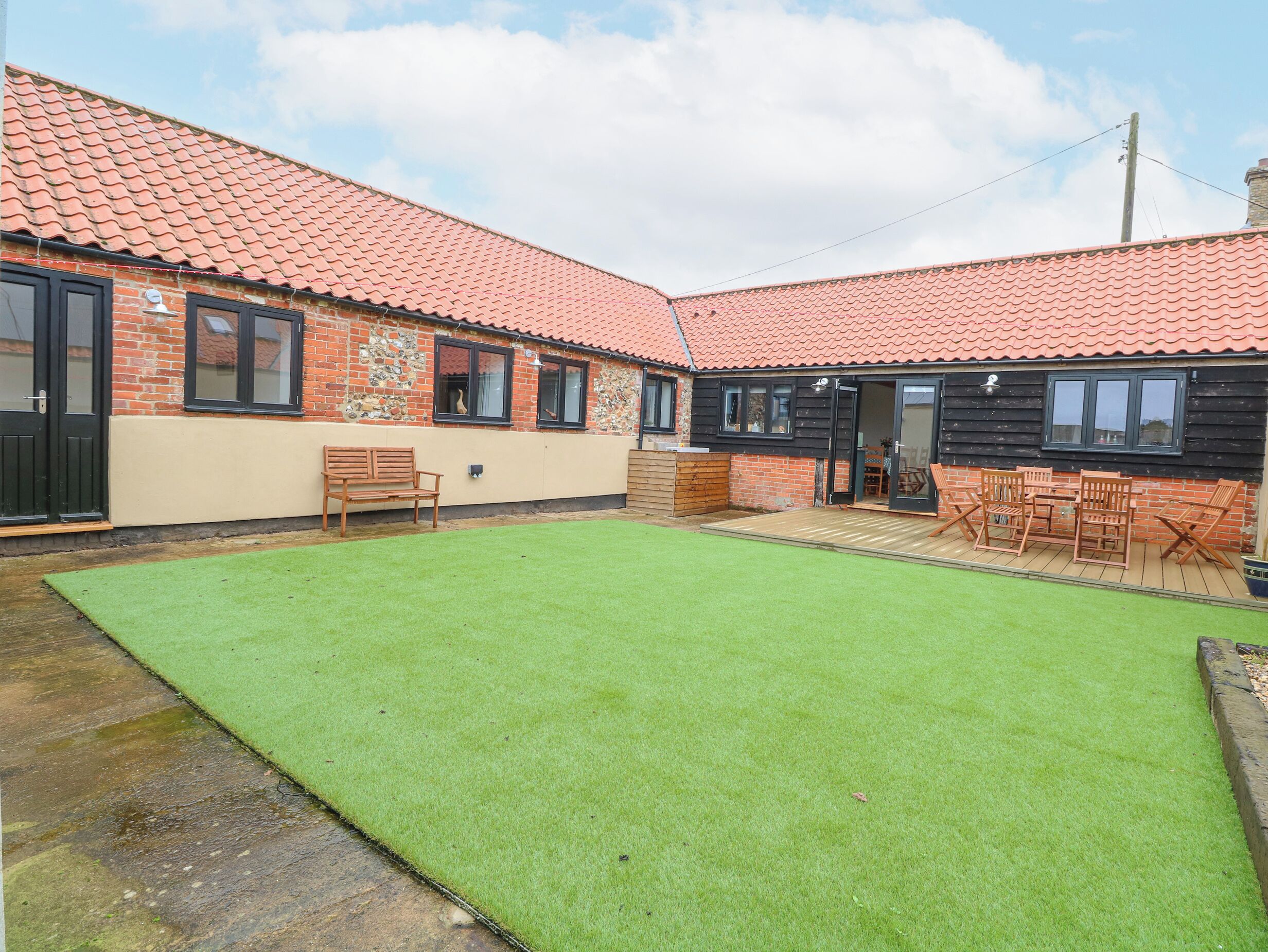 A two-story house with a red-tiled roof, a green lawn, and a patio area with wooden furniture.