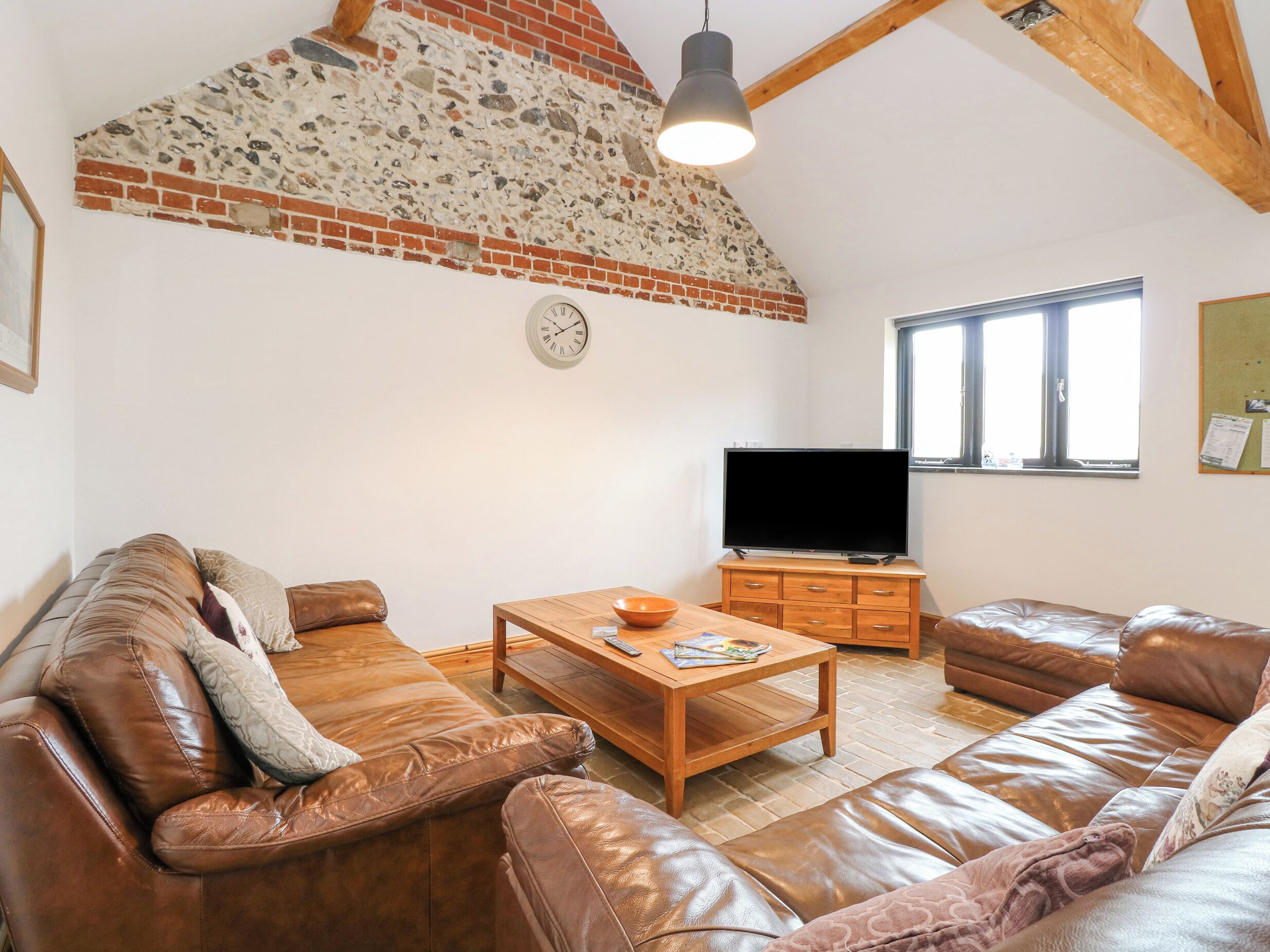 A living room with leather sofas, a wooden coffee table, a TV on a wooden stand, a clock, and a window with natural light.