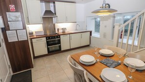 A modern kitchen with white cabinets, a stainless steel range hood, and a dining area with a wooden table set for four. - Wood Cottage (Haverfordwest)