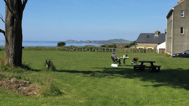 A grassy outdoor area with a picnic table and chairs, a tree, and a stone building with a view of the sea. - Wood Cottage (Haverfordwest)