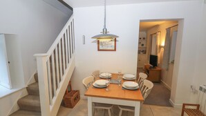 A dining area with a wooden table set for four, white chairs, a hanging pendant light, and a staircase with white railings. - Wood Cottage (Haverfordwest)