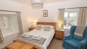 A neatly arranged bedroom with a bed, bedside tables, a wooden chest, a blue armchair, and a window with curtains. - Wood Cottage (Haverfordwest)