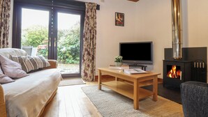A cozy living room with a sofa, a TV, a fireplace, and a large window with curtains. - Court Cottage (Looe)