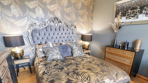 Une chambre à coucher avec une tête de lit capitonnée, une couvre-lit à motifs, deux lampes de chevet, une table de chevet, une commode et un tableau encadré au mur.