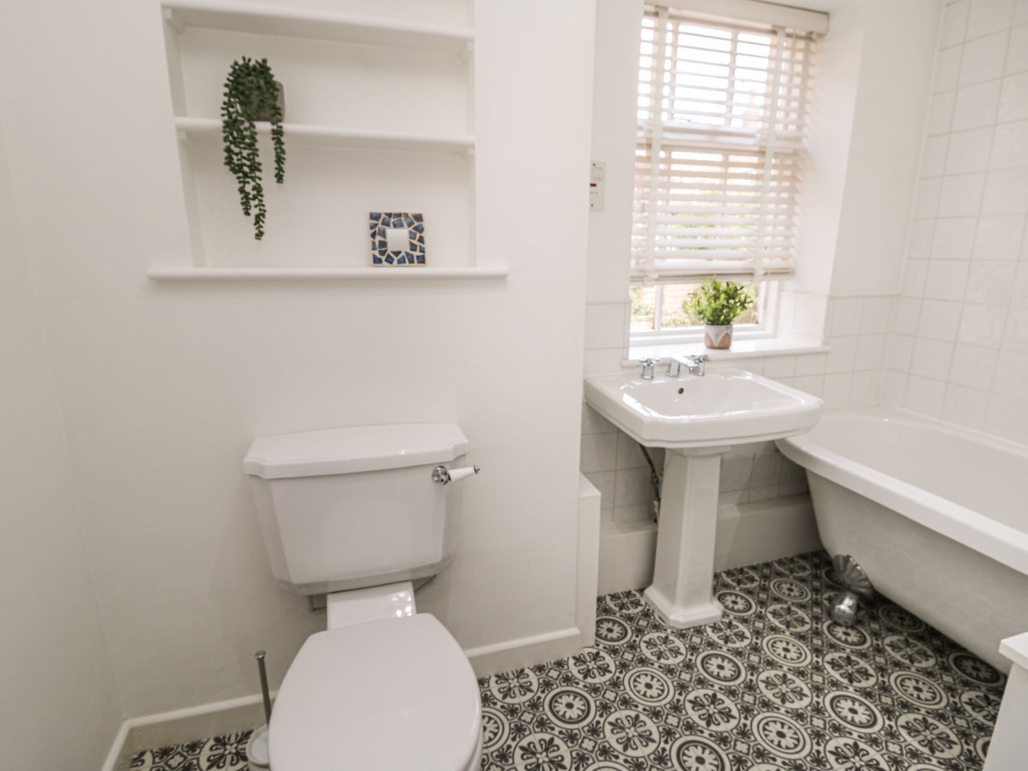 Un bagno con servizi igienici, lavabo e vasca da bagno. C'è una finestra con persiane e una pianta sul davanzale. Il pavimento presenta un motivo a piastrelle.