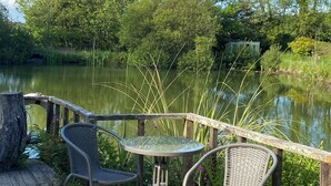 Una terraza de madera con dos sillas de mimbre y una mesa de vidrio, con vista a un estanque tranquilo rodeado de abundante vegetación.