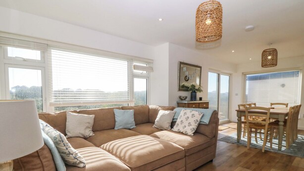 A spacious living area with a large L-shaped sectional sofa, a dining table with chairs, and a hanging woven light fixture. - Awel-y-mor (Holyhead)