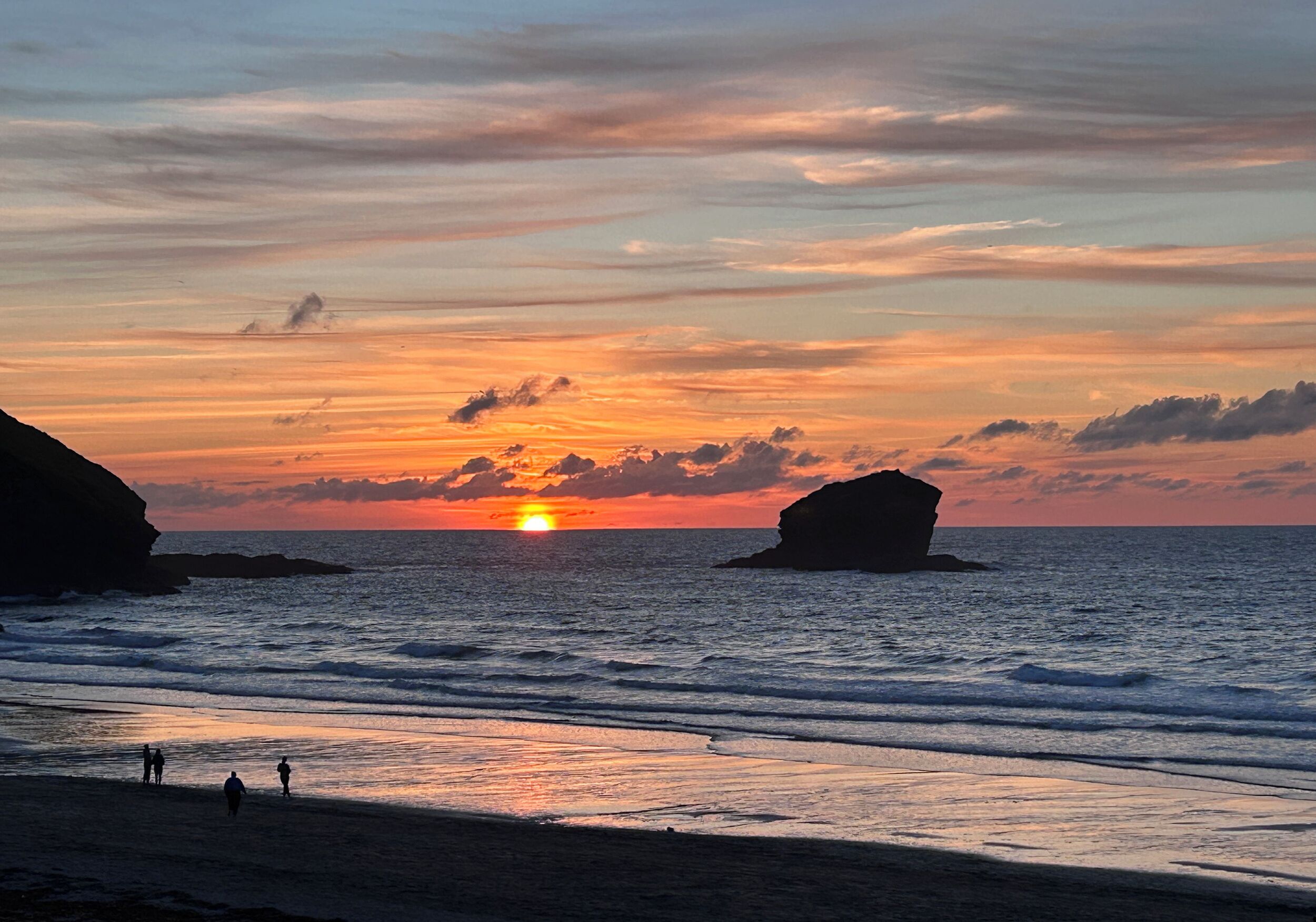 A beach scene with a sunset, waves, and a few people walking.
