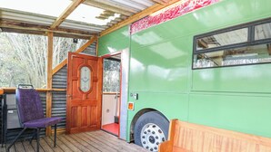 A green bus converted into a stationary living space with a wooden deck, a purple chair, a wooden bench, and a door with a stained glass window. - Walter (Wells-next-the-Sea)