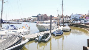 A marina with various boats docked, a wooden pier, and buildings in the background. - Walter (Wells-next-the-Sea)