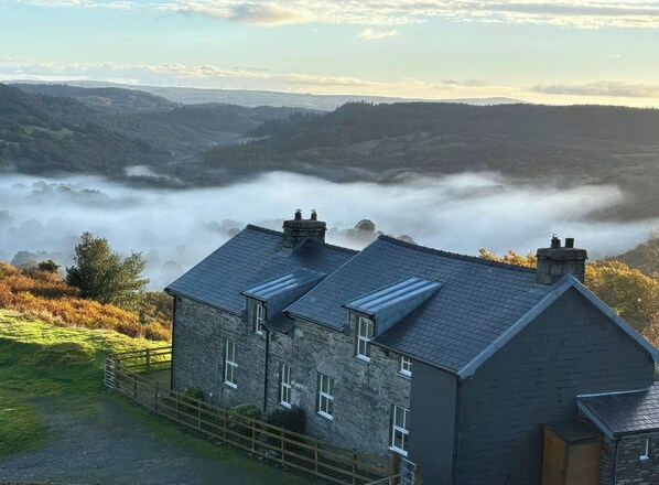 A stone building with multiple chimneys, a fenced yard, and a view of rolling hills and a valley shrouded in mist. - Ty Siabod (Betws-Y-Coed)