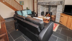 A living room with a stone fireplace, a black leather sofa, a wooden TV stand, and a staircase. - Ty Siabod (Betws-Y-Coed)