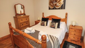 A neatly made bed with a wooden headboard, a bedside table with a lamp, a dresser with a mirror, and a painting of a flower on the wall. - Ty Siabod (Betws-Y-Coed)