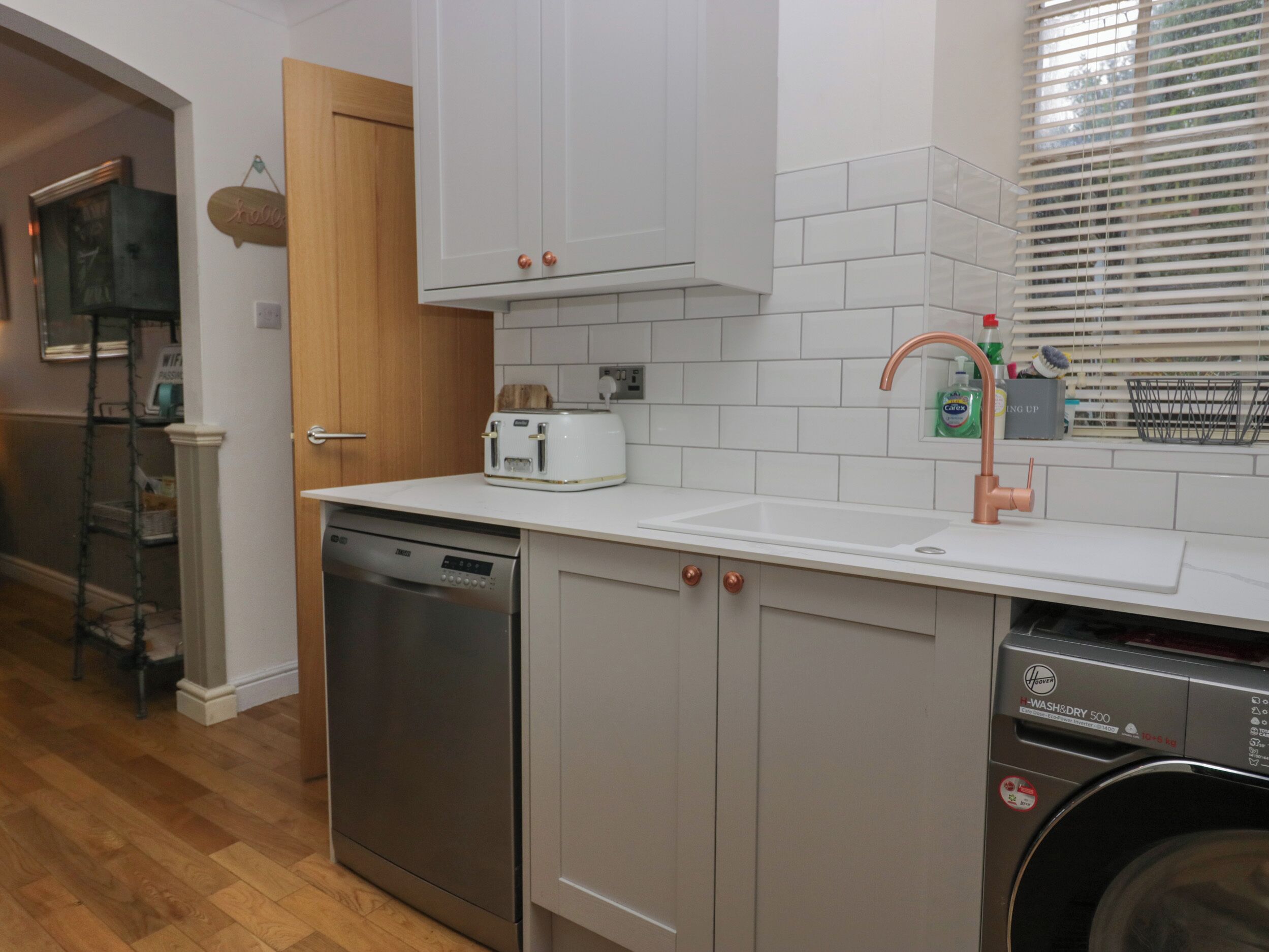 A modern kitchen with white cabinetry, a stainless steel dishwasher, a built-in oven, a white countertop, and a sink with a copper faucet.