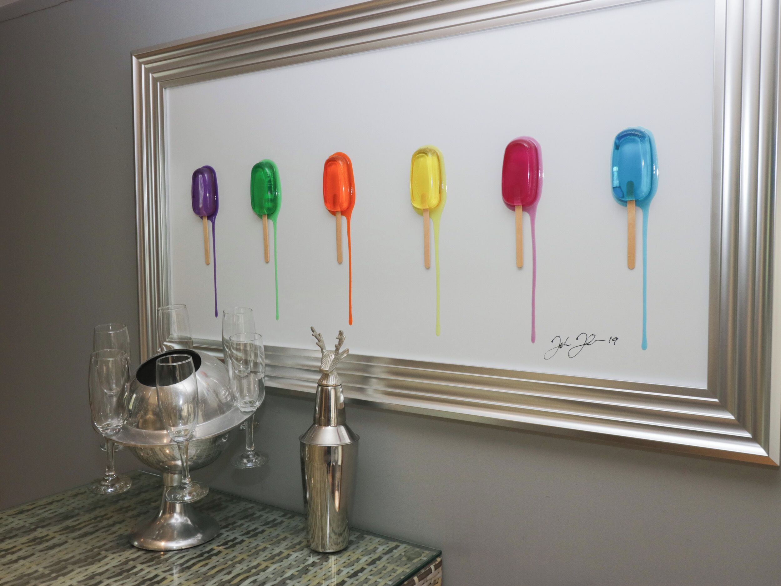 A framed artwork of colorful popsicles on a wall, a silver cocktail shaker, and a silver goblet with glasses on a table.