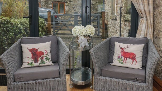 Two wicker chairs with floral cushions, a metal basket, and a glass table in a room with stone walls and a view of a stone building. - 1 Boarbank Farm (Grange-over-Sands)