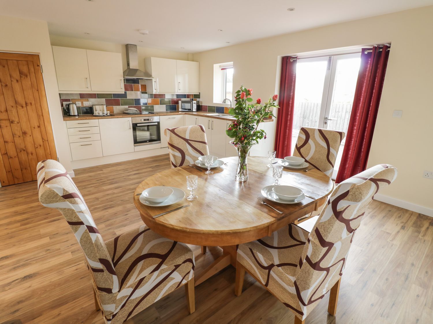 A dining area with a round wooden table, six patterned chairs, a vase with flowers, and a kitchen in the background.
