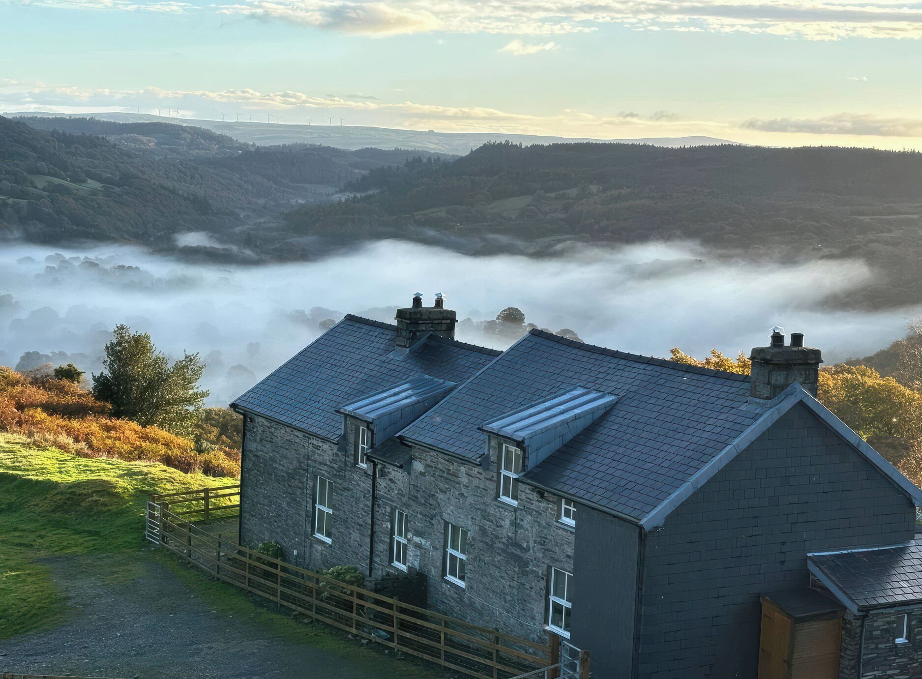 A stone building with multiple chimneys, a fenced yard, and a view of rolling hills and a valley shrouded in mist.