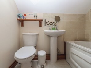 Een kleine badkamer met toilet, wastafel en bad. Boven het toilet bevindt zich een plankje met toiletartikelen.