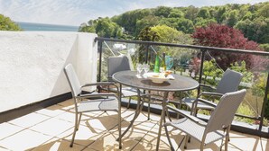 A balcony with a round table and four wicker chairs, overlooking a lush green landscape and a body of water. - Valley View, 9 St Elmo Court (Salcombe)