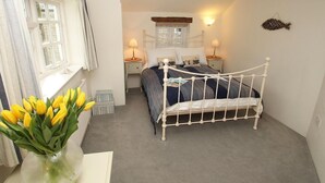 Une chambre à coucher avec un lit en métal blanc, une fenêtre avec des rideaux, une table de chevet avec une lampe et un vase rempli de fleurs jaunes.