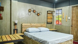 A bedroom with a wooden bed, a small table, a bench, a wall-mounted lamp, and a window with a colorful view. - Bhive Hotel (Iloilo)