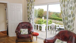 A room with a wooden floor, a brown leather armchair with a leaf-patterned pillow, and a view of a garden through a glass door. - Lakeview Lodge (Foxford)