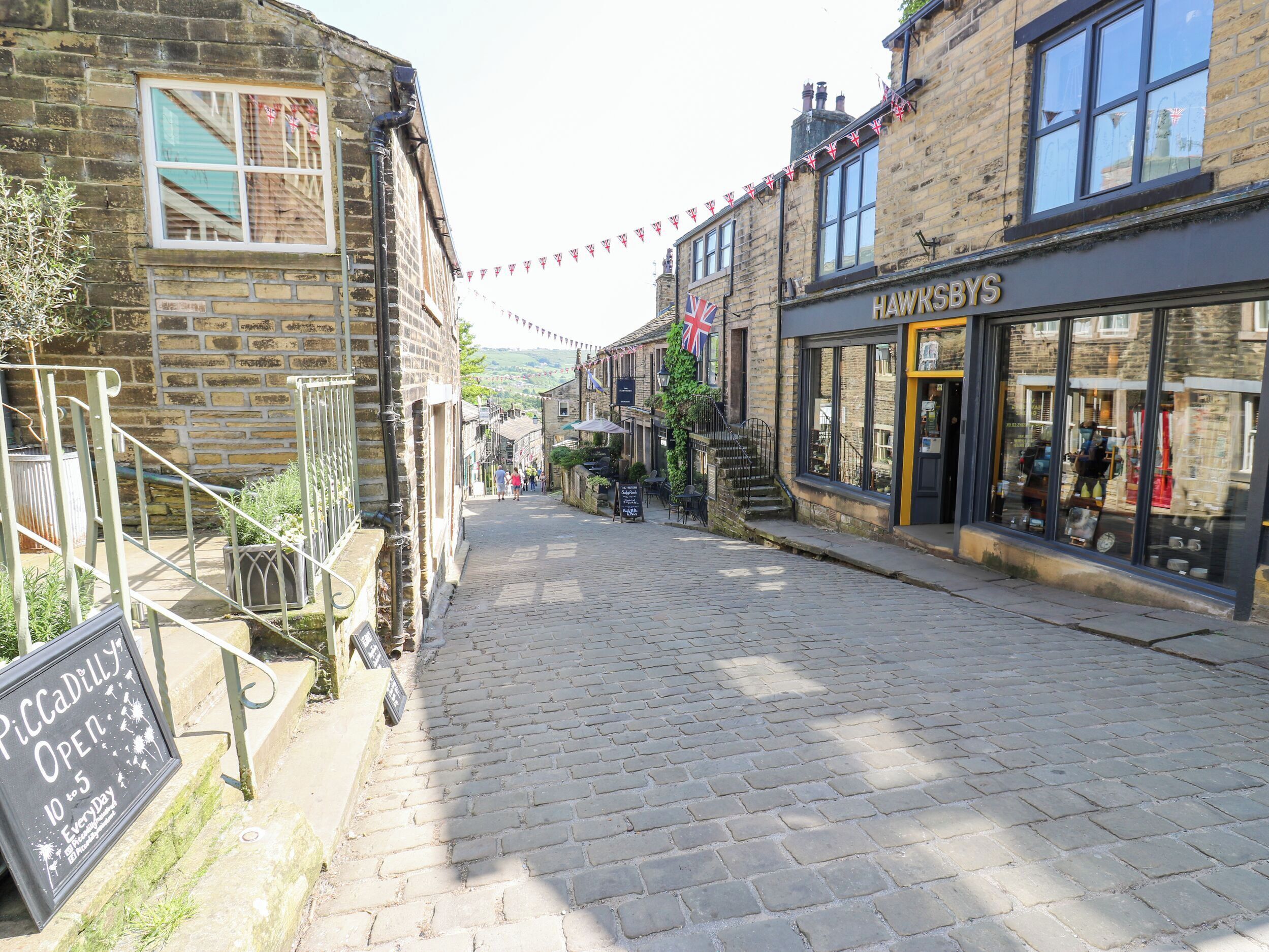 A cobblestone street with buildings on either side, including a shop named 'Hawksbys'. There is a sign for 'Piccadilly Open 10 to 5 Every Day'.