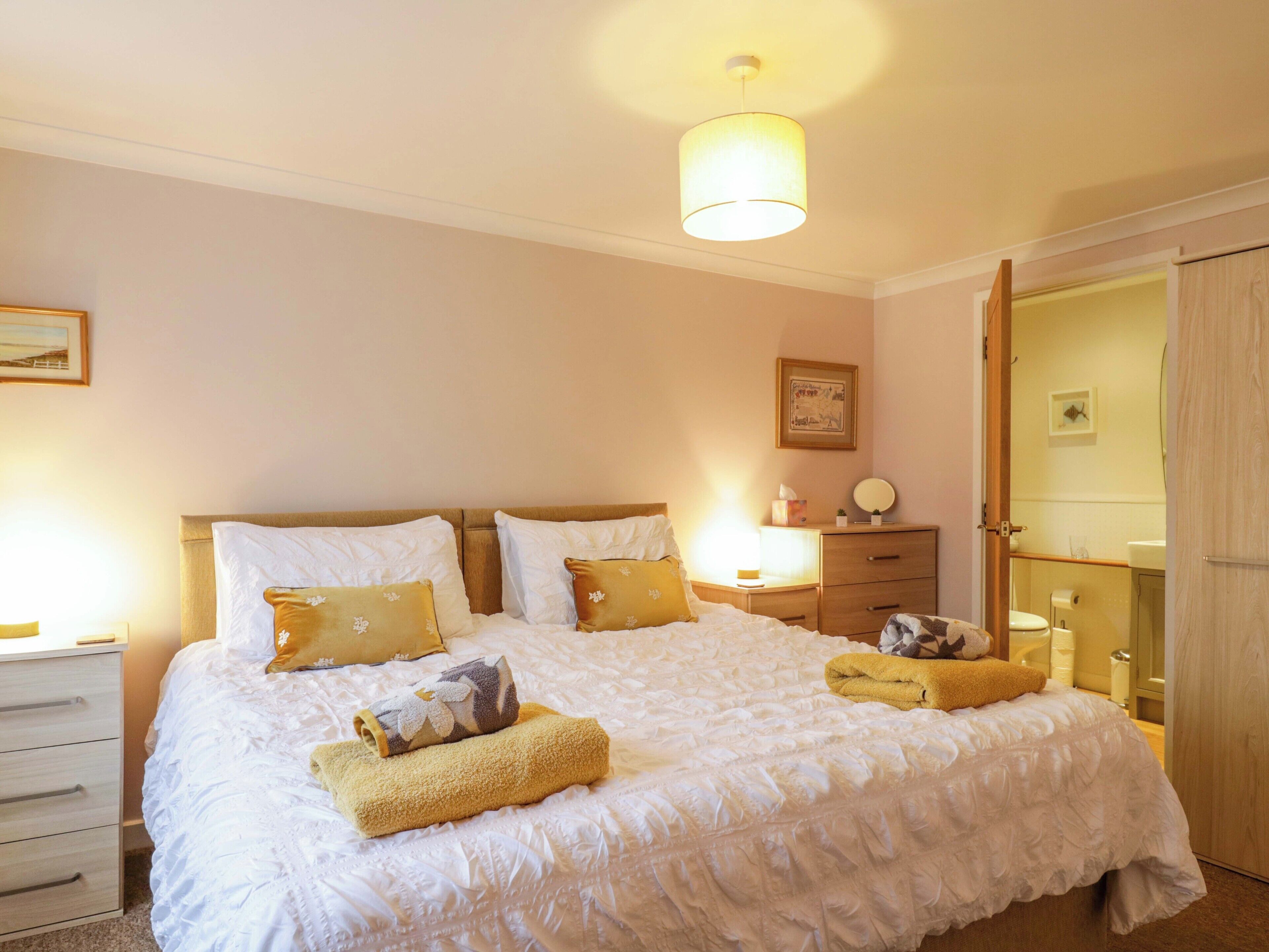 Una cama bien tendida con ropa de cama blanca y dos almohadas decorativas, una mesita de noche con lámpara y un armario de madera.
