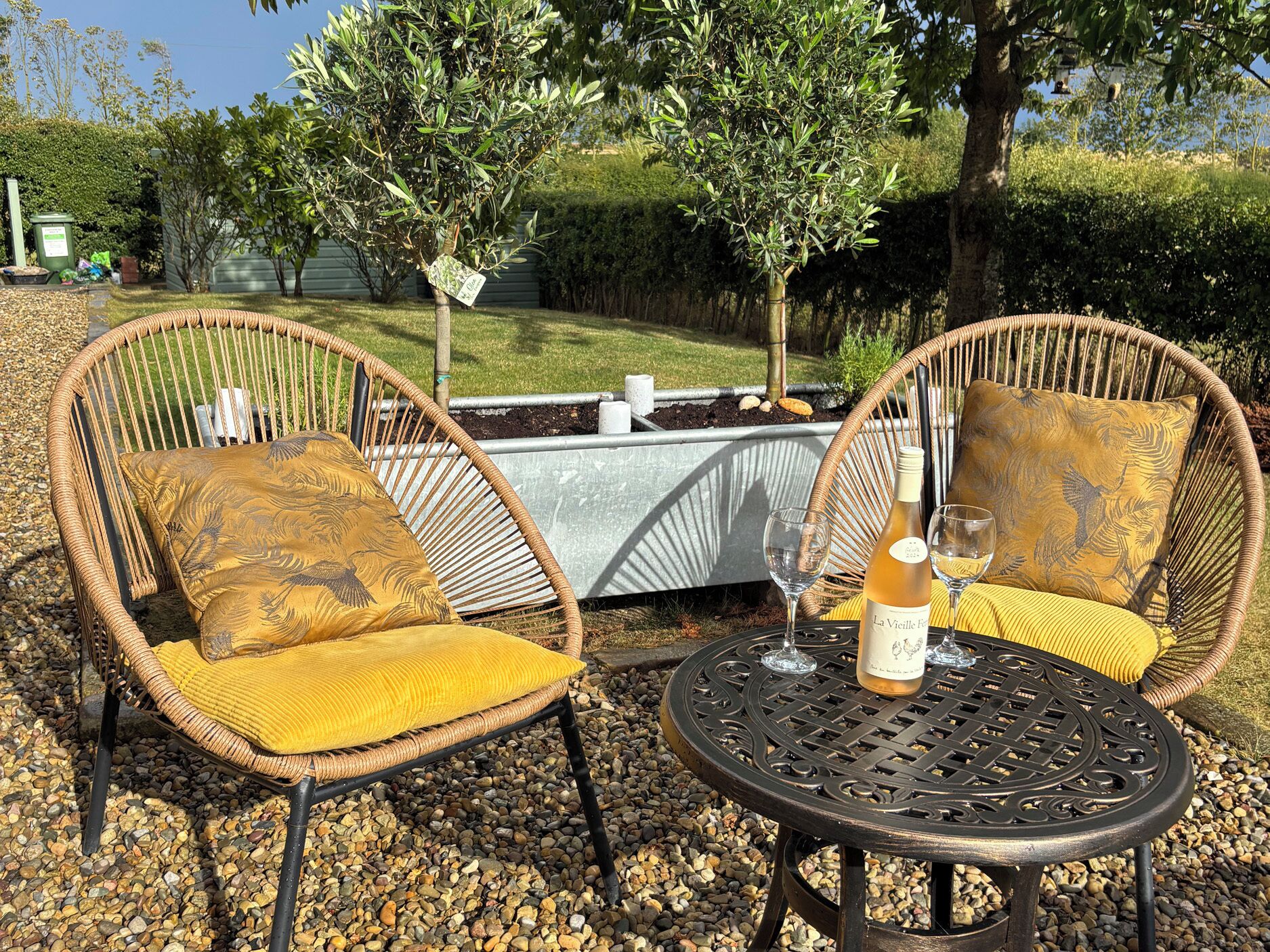 Two wicker chairs with yellow cushions, a round metal table, and a bottle of wine.