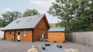 A wooden cabin with a sloped roof, a small attached shed, and a gravel driveway with large rocks. - Bradan Lodge (Dalmally)