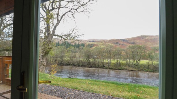 A view from a room with a wooden deck overlooking a river, surrounded by hills and trees. - Bradan Lodge (Dalmally)