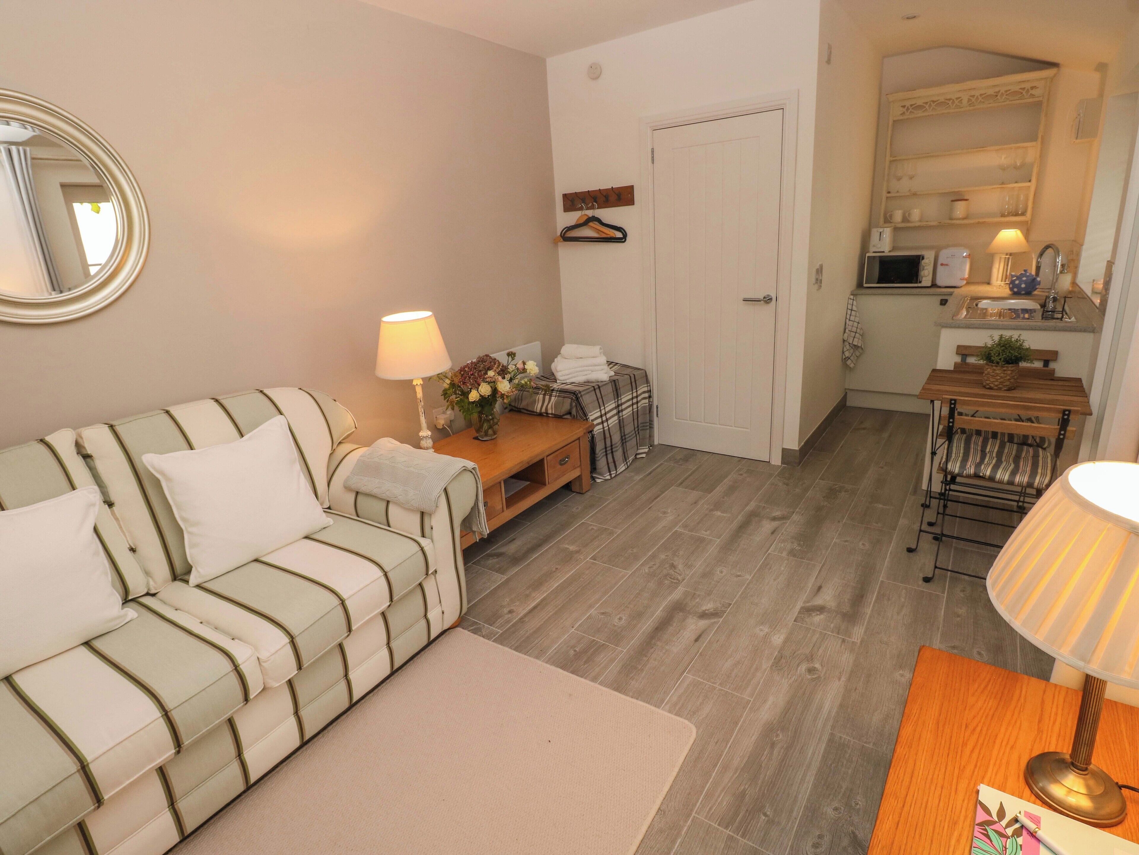 A cozy living room with a striped sofa, a small wooden table with a lamp, and a door leading to a kitchen area.