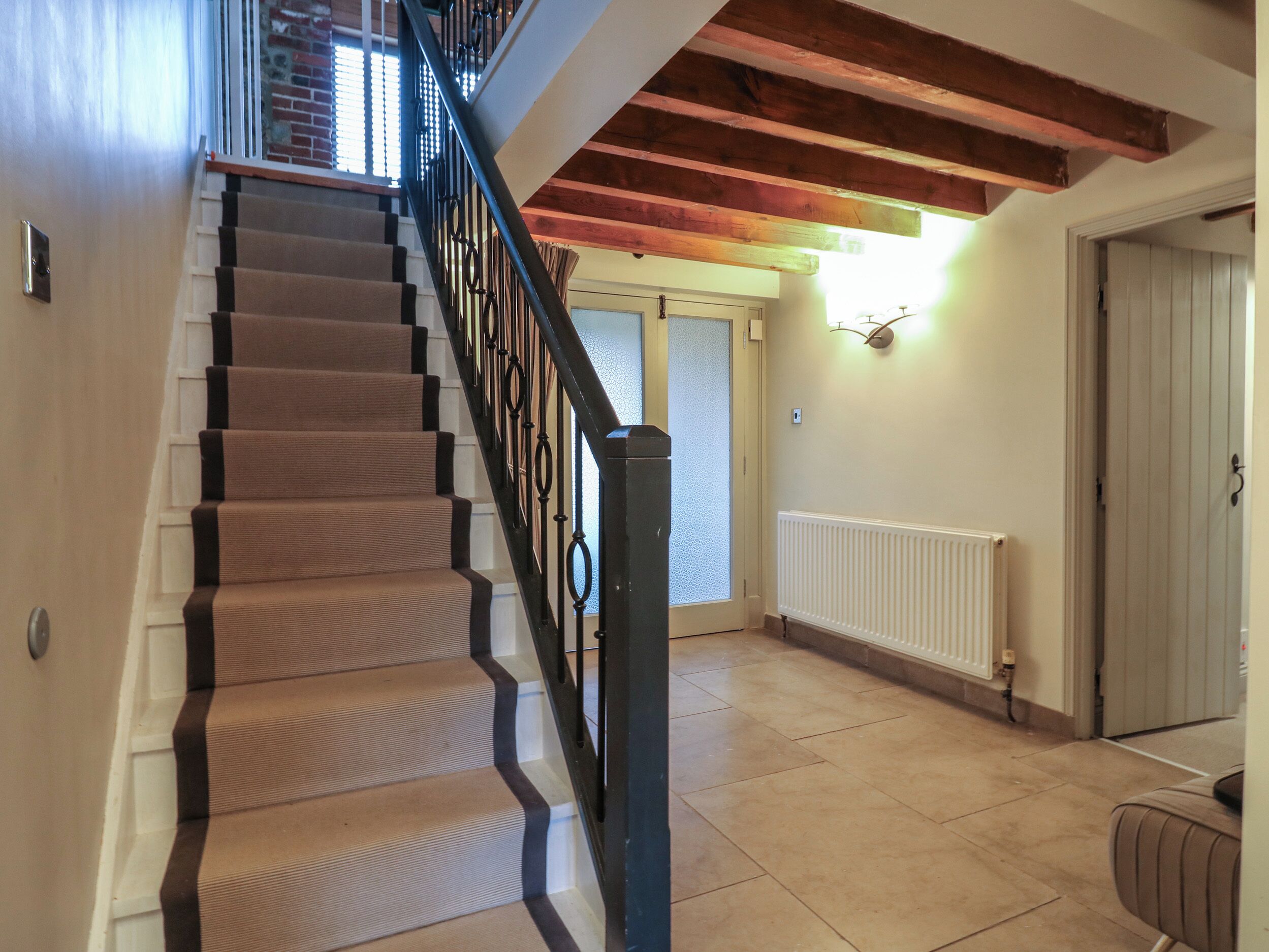 Una escalera con peldaños cubiertos de alfombra beige y barandillas negras, que conduce a una habitación con una puerta blanca y un radiador en la pared.