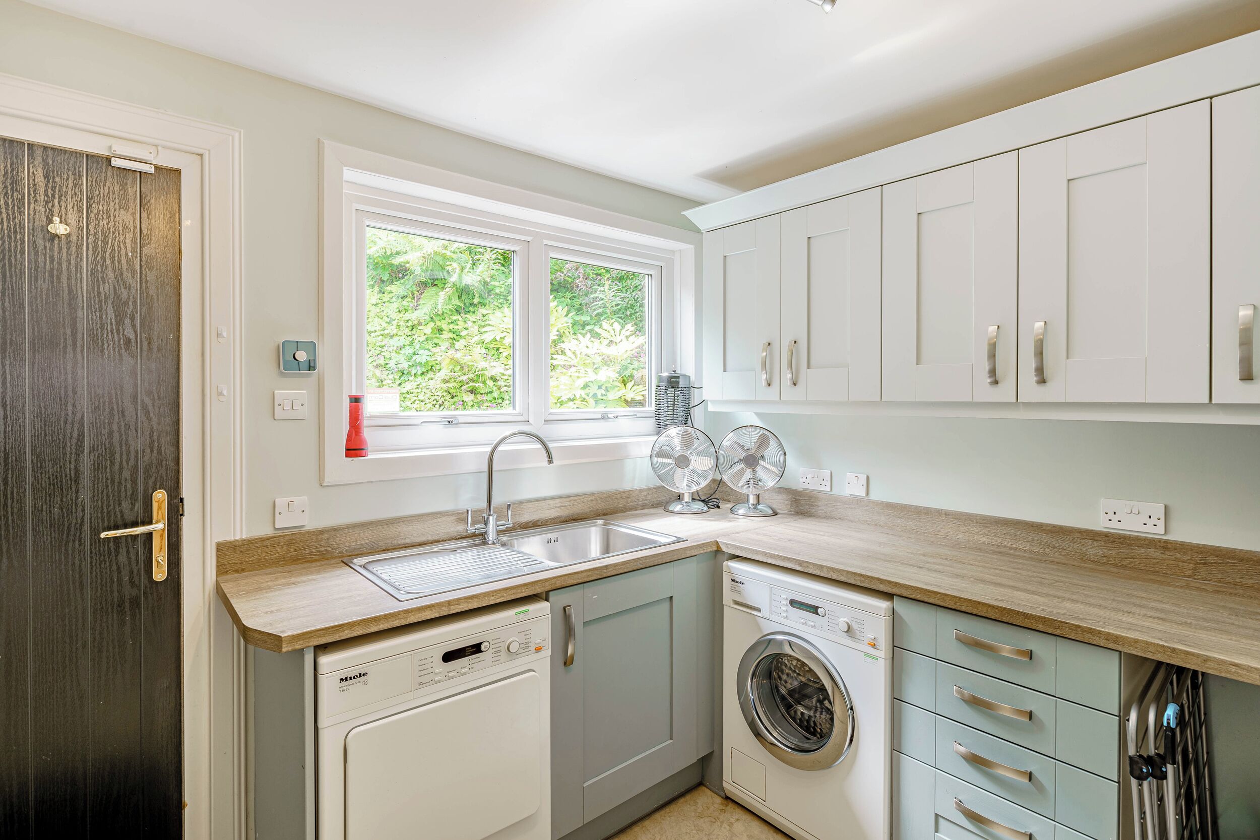 A modern kitchen with a dishwasher, washing machine, and a window with a view of greenery.